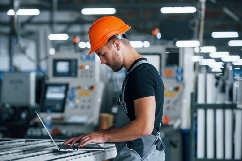 Typing on keyboard. Industrial worker indoors in factory. Young technician with orange hard hat